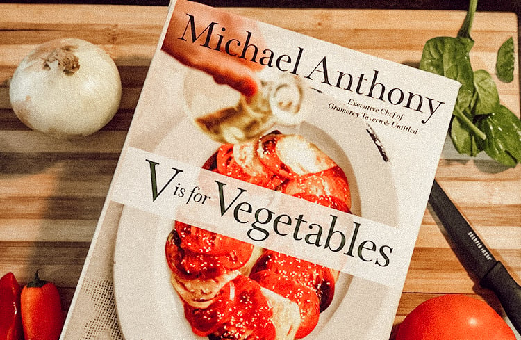 A vegetable cookbook on a cutting board with a knife and fresh vegetables creatively laid out.
